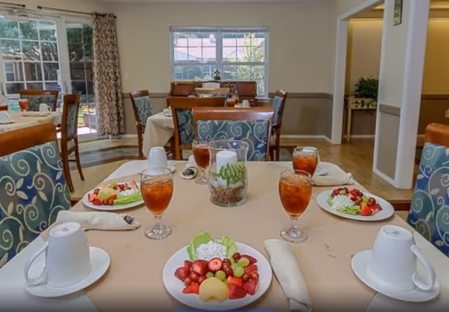 A dining room in a senior living facility with tables set for a meal. Each table has plates with fruit and salad, glasses of iced tea, white coffee cups, and napkins. The room has large windows letting in natural light, patterned chairs, and a view of an outdoor area through glass doors.