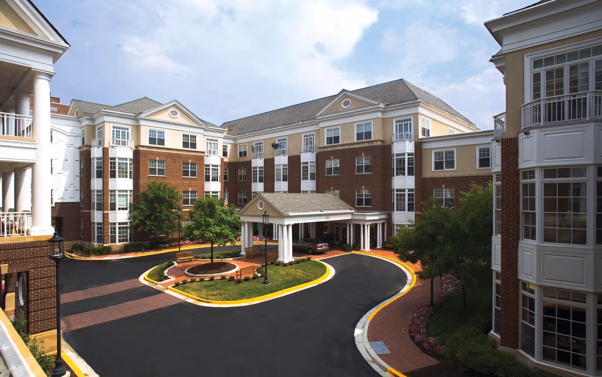 Exterior view of a multi-story assisted living facility with a circular driveway, landscaped greenery, benches, and a covered entrance under a partly cloudy sky.