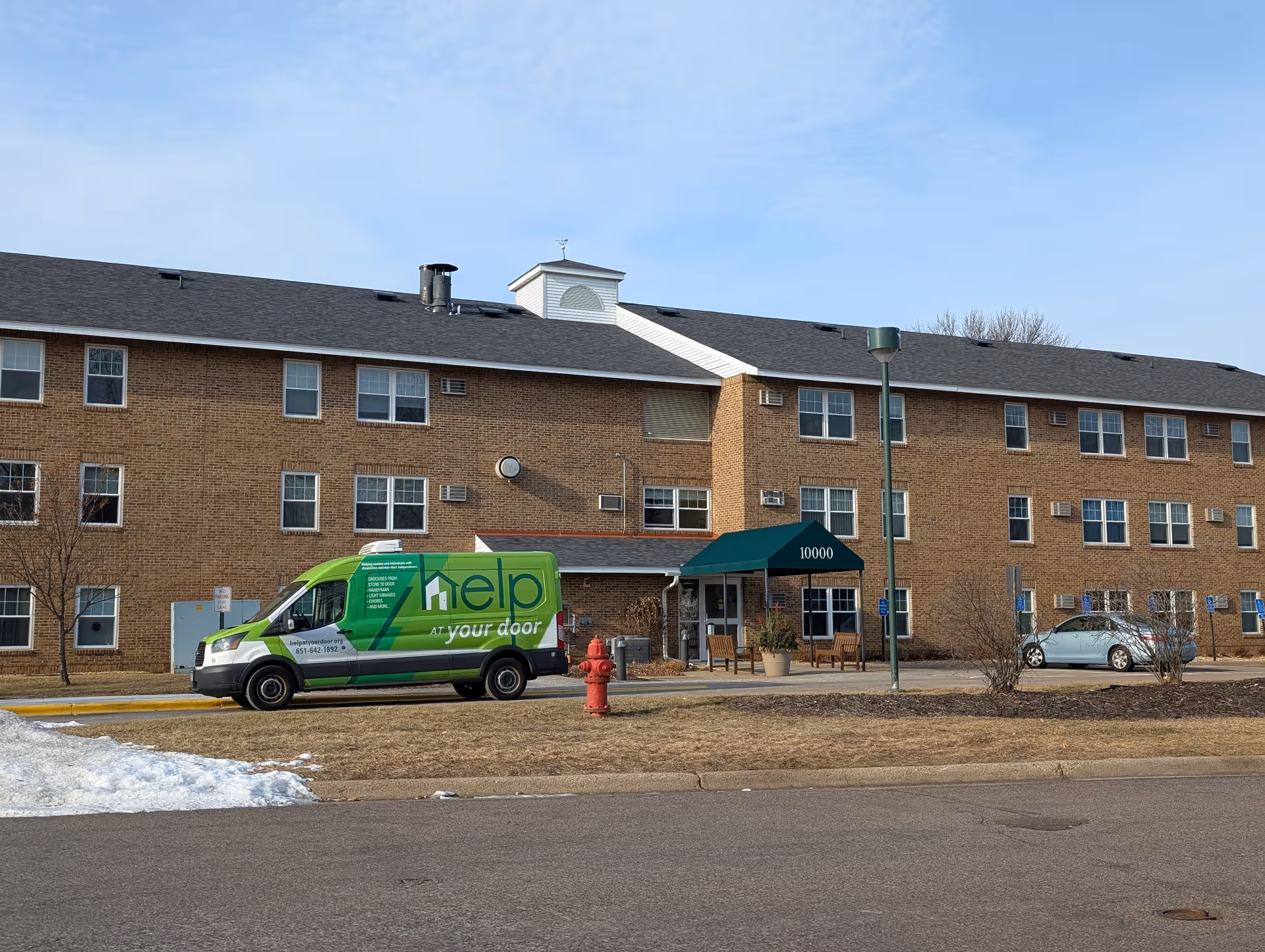 Exterior view of a three-story brick building with multiple windows and air conditioning units. A green van with the text 'help at your door' is parked in front near a red fire hydrant. There is a green awning over the entrance with the number 10000 displayed. The ground is partly covered with snow and dry grass under a clear blue sky.