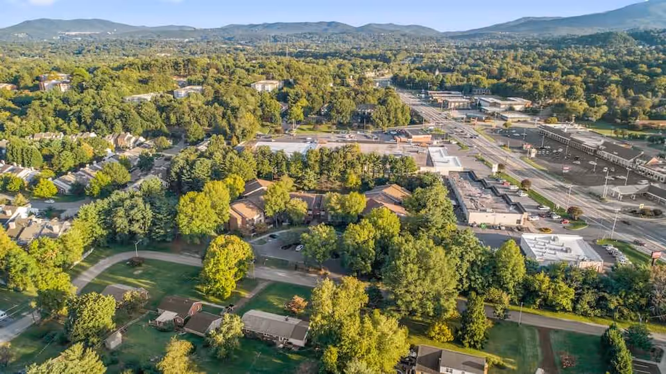 Aerial view of a tree-lined suburban neighborhood and commercial strip with roads and distant mountains.