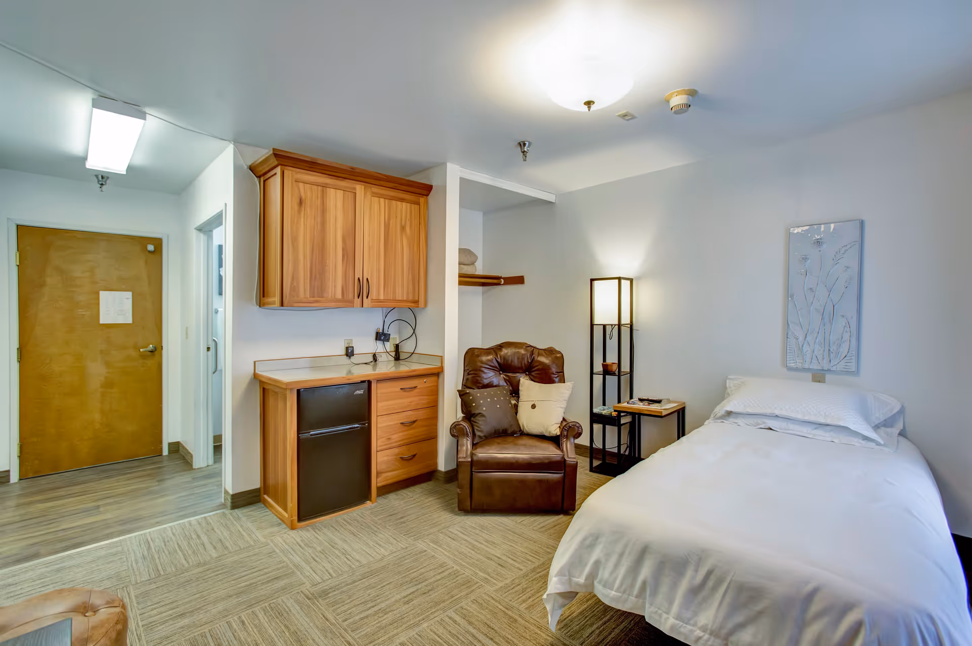 A senior living facility bedroom featuring a single bed with white bedding, a brown leather armchair with two pillows, a tall floor lamp, a small side table, wooden cabinets with a mini fridge underneath, and a wooden door leading to another room. The room has light-colored walls and carpeted flooring.