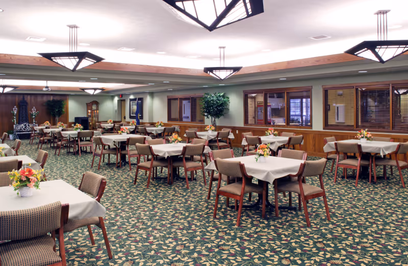 A spacious dining room with multiple tables covered in white tablecloths, each adorned with a small floral centerpiece. The room features patterned carpet flooring, wooden paneling on the walls, large windows with blinds, and modern geometric ceiling lights.