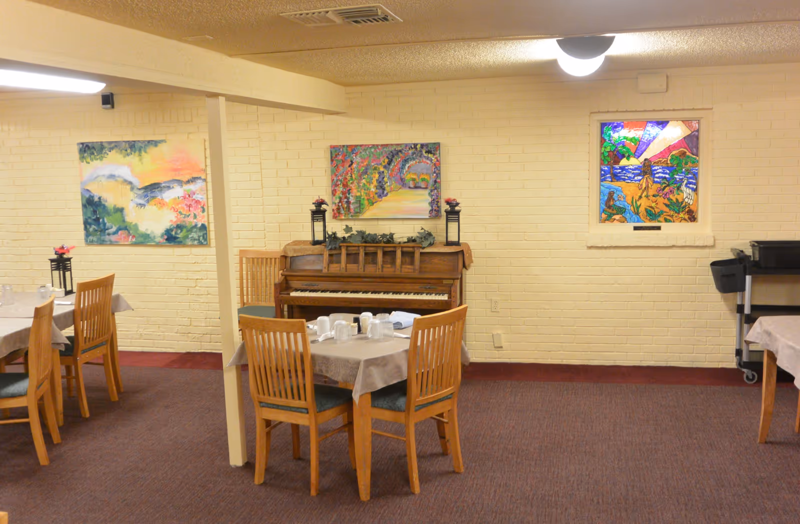 Dining room with set tables and wooden chairs, a piano against a painted brick wall, and colorful artwork.