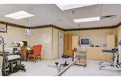 Rehabilitation room with exercise equipment, therapy chairs, a TV, and storage cabinets.