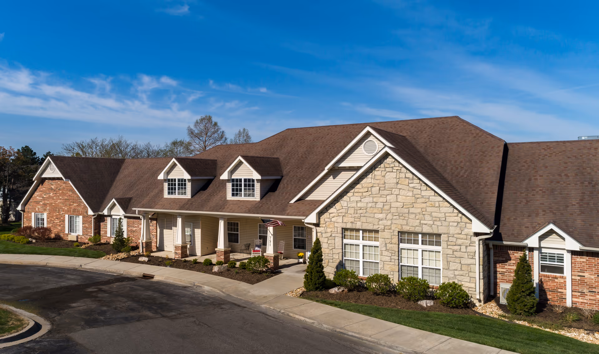 Exterior view of a single-story senior living facility building with a combination of brick and stone facade, multiple windows, a brown shingled roof, and a small covered porch with an American flag. The building is surrounded by landscaped bushes and a paved driveway under a clear blue sky.