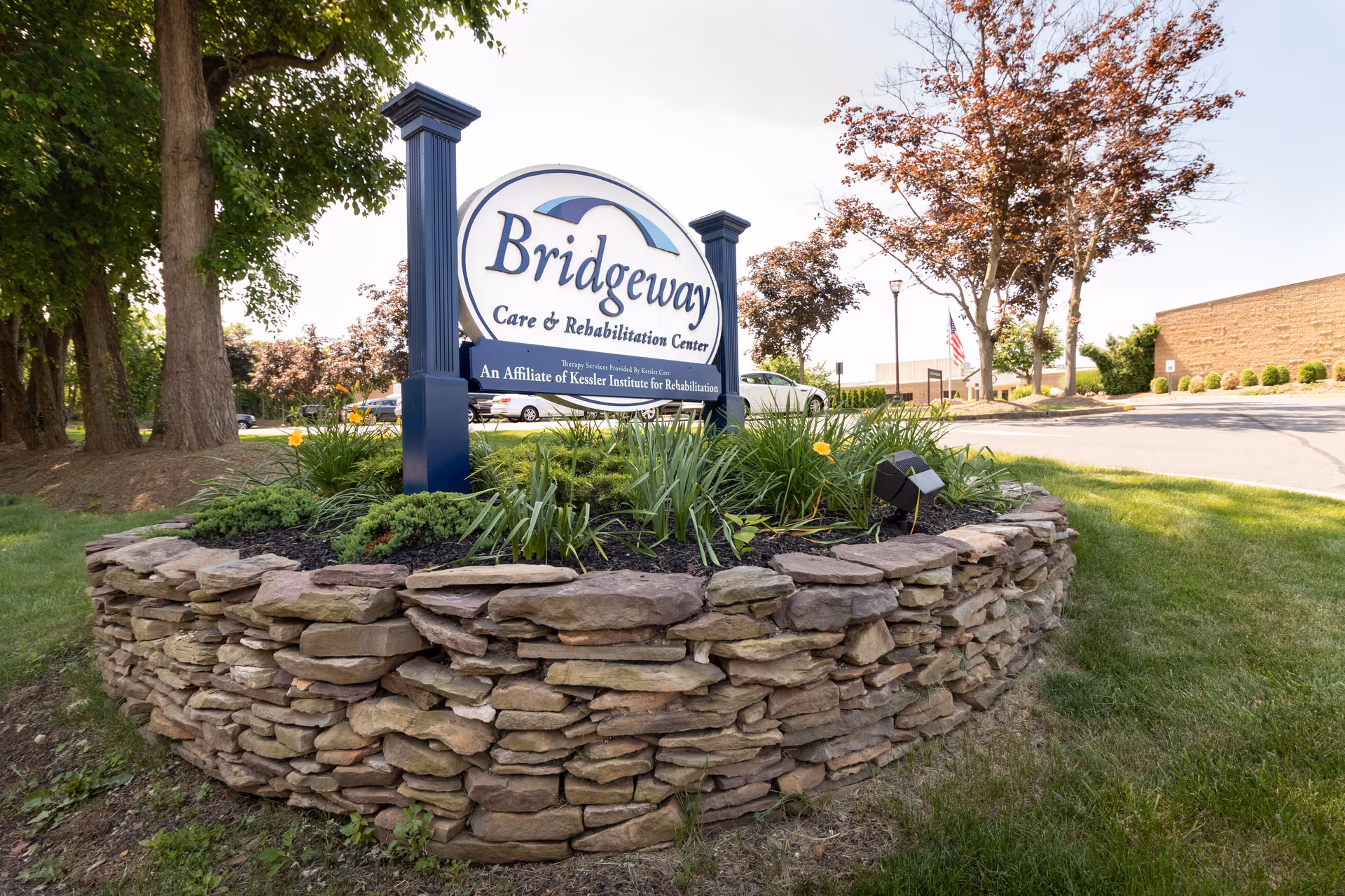 Outdoor view of a stone-bordered garden bed with a sign that reads 'Bridgeway Care & Rehabilitation Center, An Affiliate of Kessler Institute for Rehabilitation' surrounded by trees, grass, and a parking lot in the background.