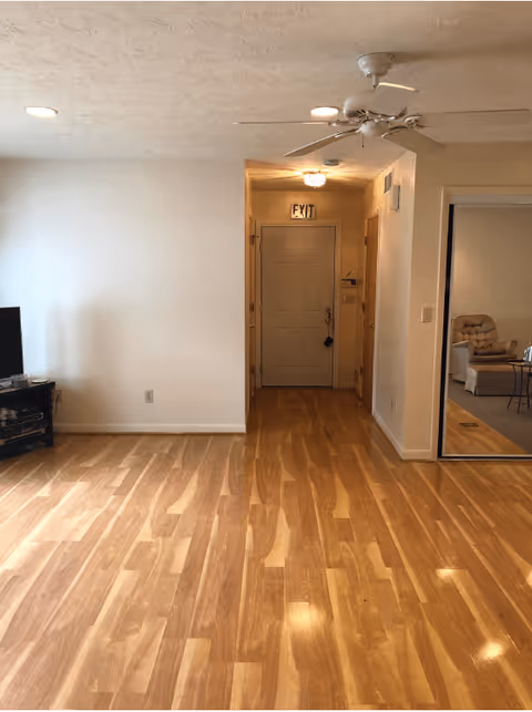 Interior view of a room with wooden flooring, a ceiling fan, and recessed lighting. There is a hallway leading to a door with an exit sign above it. To the right, there is a mirrored sliding door reflecting a seating area with a cushioned chair and a small table. On the left side, a TV is placed on a stand against the wall.