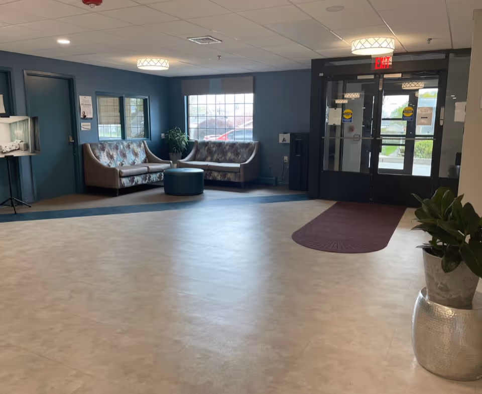 Interior view of a facility lobby area with two patterned sofas, a round ottoman, a large window, double glass doors with exit signs, a potted plant in a silver pot, and a water dispenser against a blue wall.