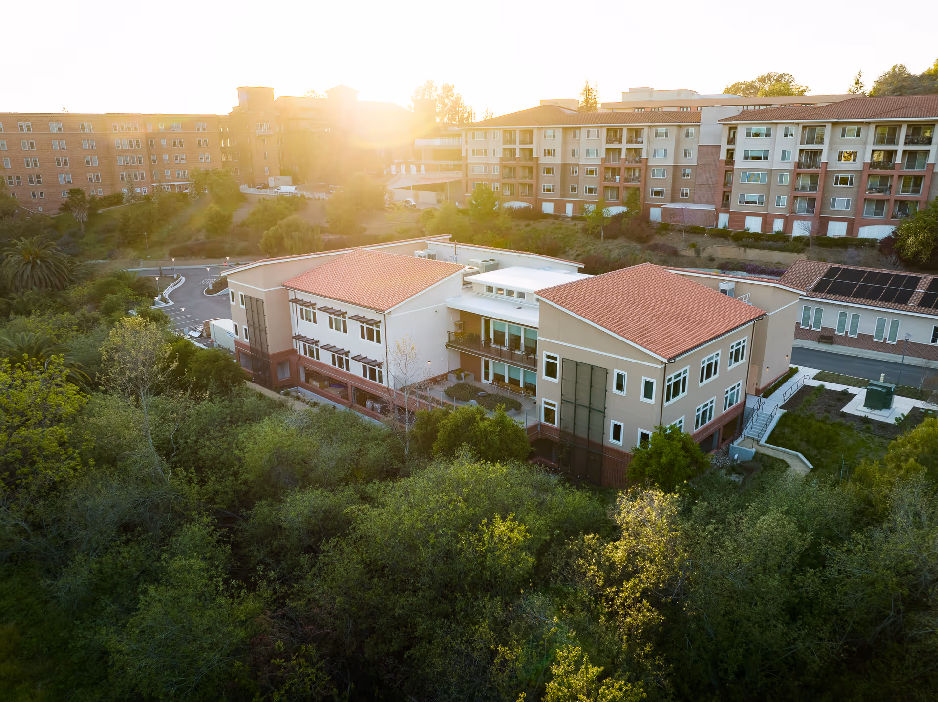 Aerial view of a senior living facility with multiple buildings featuring red-tiled roofs surrounded by greenery and trees, with the sun shining brightly in the background.