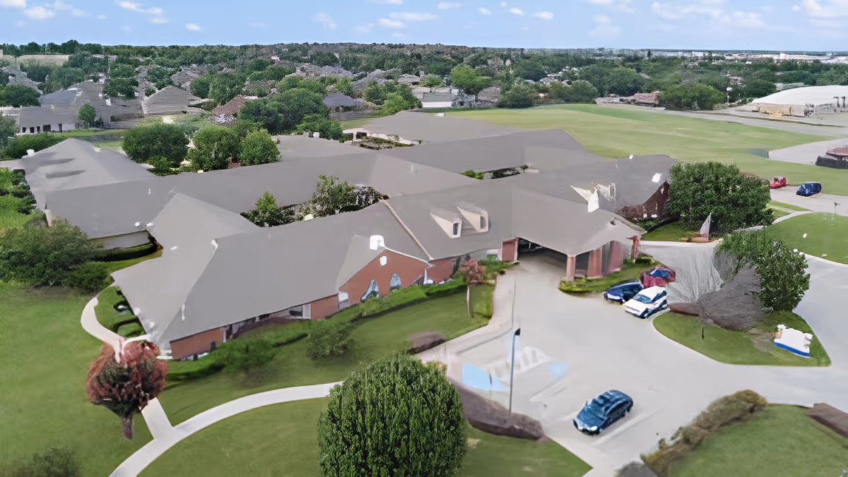 Aerial view of a senior living facility building with a large parking area, surrounded by green lawns and trees under a blue sky.