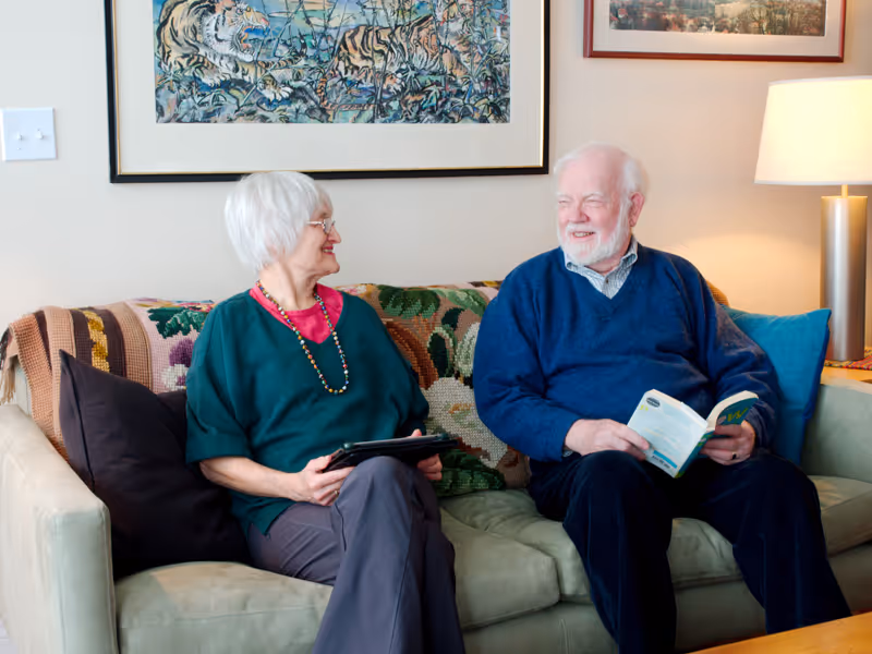 An elderly woman and an elderly man sitting on a couch in a living room. The woman is holding a tablet and smiling at the man, who is holding a book and smiling back. Behind them are framed artworks on the wall and a lit table lamp on a side table.