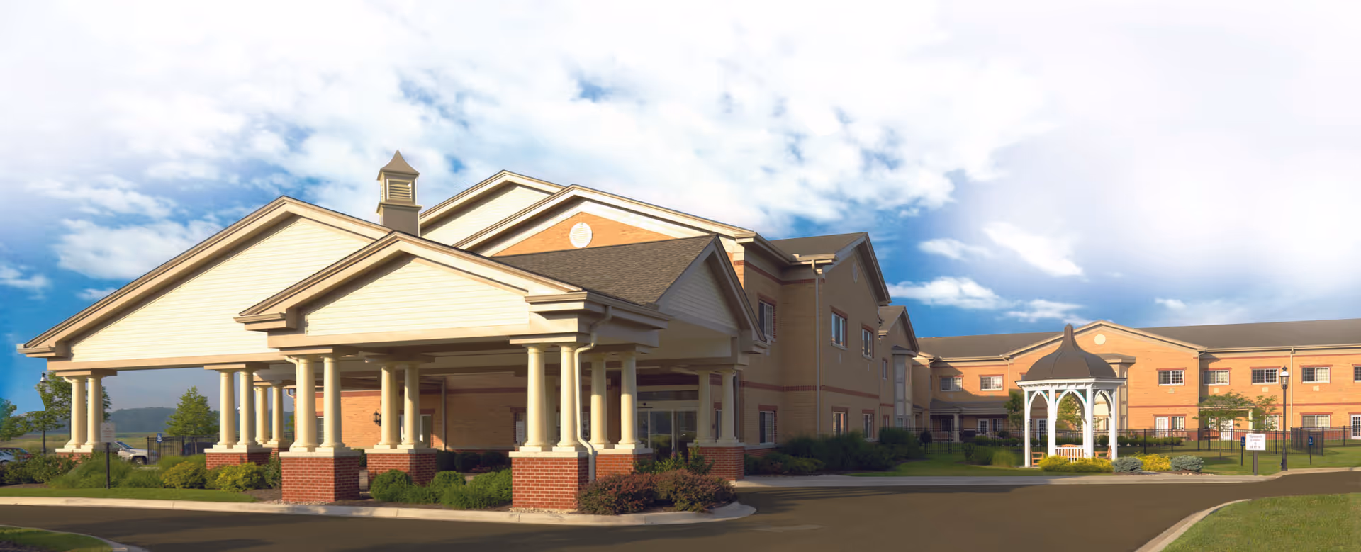 Exterior view of Stonespring Skilled Nursing Facility showing a large brick building with a covered entrance supported by columns, a gazebo on the lawn, and a partly cloudy sky above.