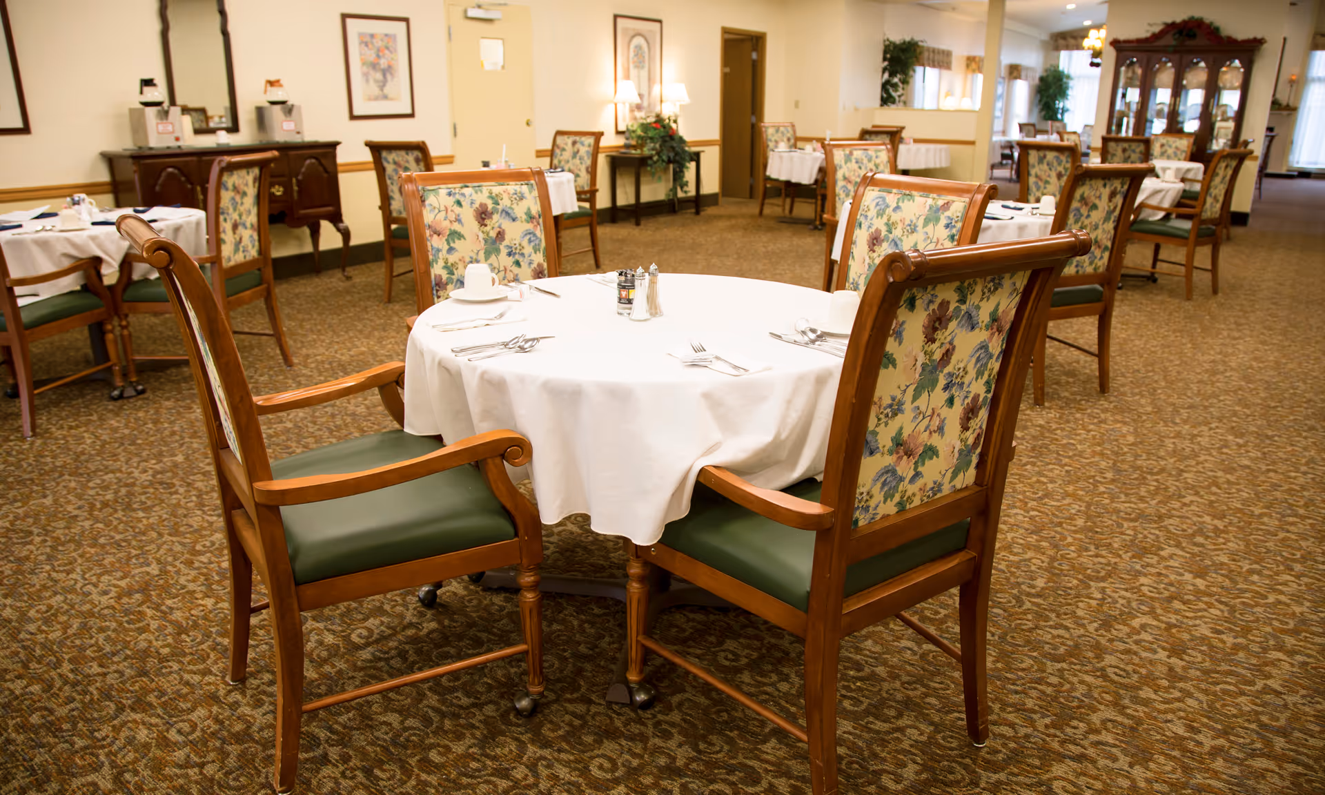 A round table set with a white tablecloth and place settings surrounded by wooden floral-upholstered chairs in a carpeted dining room.