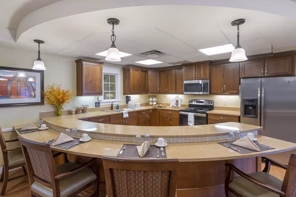 A spacious kitchen area with a large curved countertop and bar seating. The kitchen features wooden cabinets, stainless steel appliances including a refrigerator, microwave, and stove. There are pendant lights hanging from the ceiling, a window above the sink, and a decorative vase with flowers on the counter. Each seat at the bar has a placemat, napkin, cup, and utensils set up.