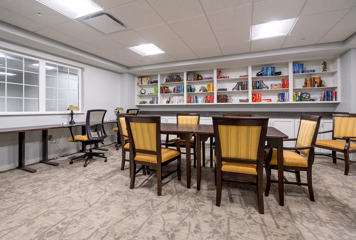 A bright common room with a central table surrounded by yellow-upholstered chairs, bookshelves along the back wall, and desks by a window.