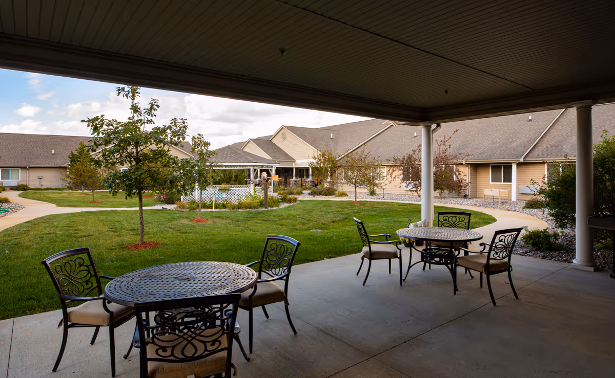 Covered patio with round metal tables and chairs overlooking a landscaped courtyard and single-story senior living buildings.