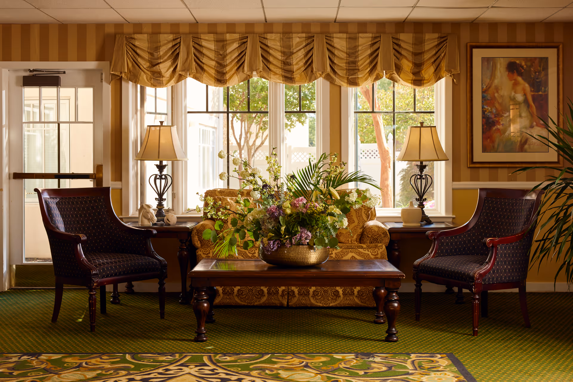 A cozy sitting area in a senior living facility featuring a patterned sofa with a large floral arrangement on a wooden coffee table in front. Two dark upholstered armchairs flank the coffee table. There are two table lamps on side tables next to the sofa, and a large window with a striped valance behind the sofa lets in natural light. A framed painting hangs on the wall to the right, and the room has warm yellow striped wallpaper and a green patterned carpet.