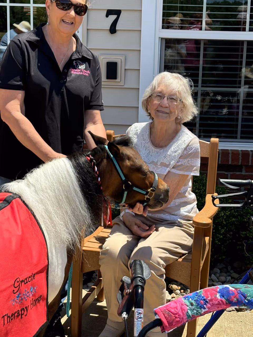 An elderly woman sitting on a wooden chair outside a building, smiling and petting a small therapy horse wearing a red blanket with the words 'Gracie Therapy Horse'. A woman standing next to her is holding the horse's reins. There is a walker with a colorful floral cover in front of the elderly woman.