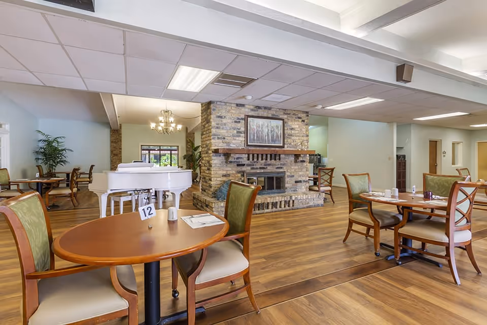 A spacious dining room with wooden floors and several round tables with chairs. A white grand piano is positioned near a brick fireplace with a framed picture above it. The room is well-lit with ceiling lights and a chandelier, and there are some plants and additional seating in the background.