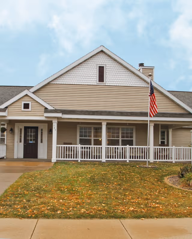 Front exterior view of a single-story residential building with beige siding, white trim, a covered porch with white railing, an American flag on a flagpole, and a lawn with some fallen leaves.