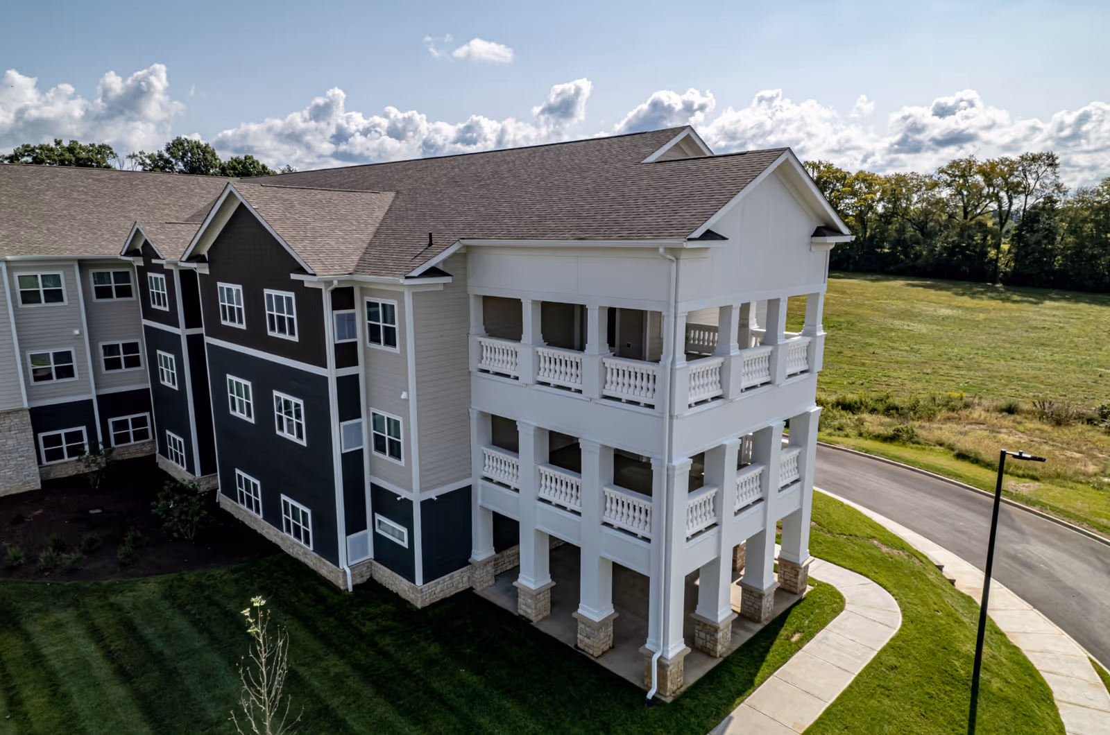Exterior view of a multi-story senior living facility building with a large covered balcony area, surrounded by green grass and a paved driveway under a partly cloudy sky.