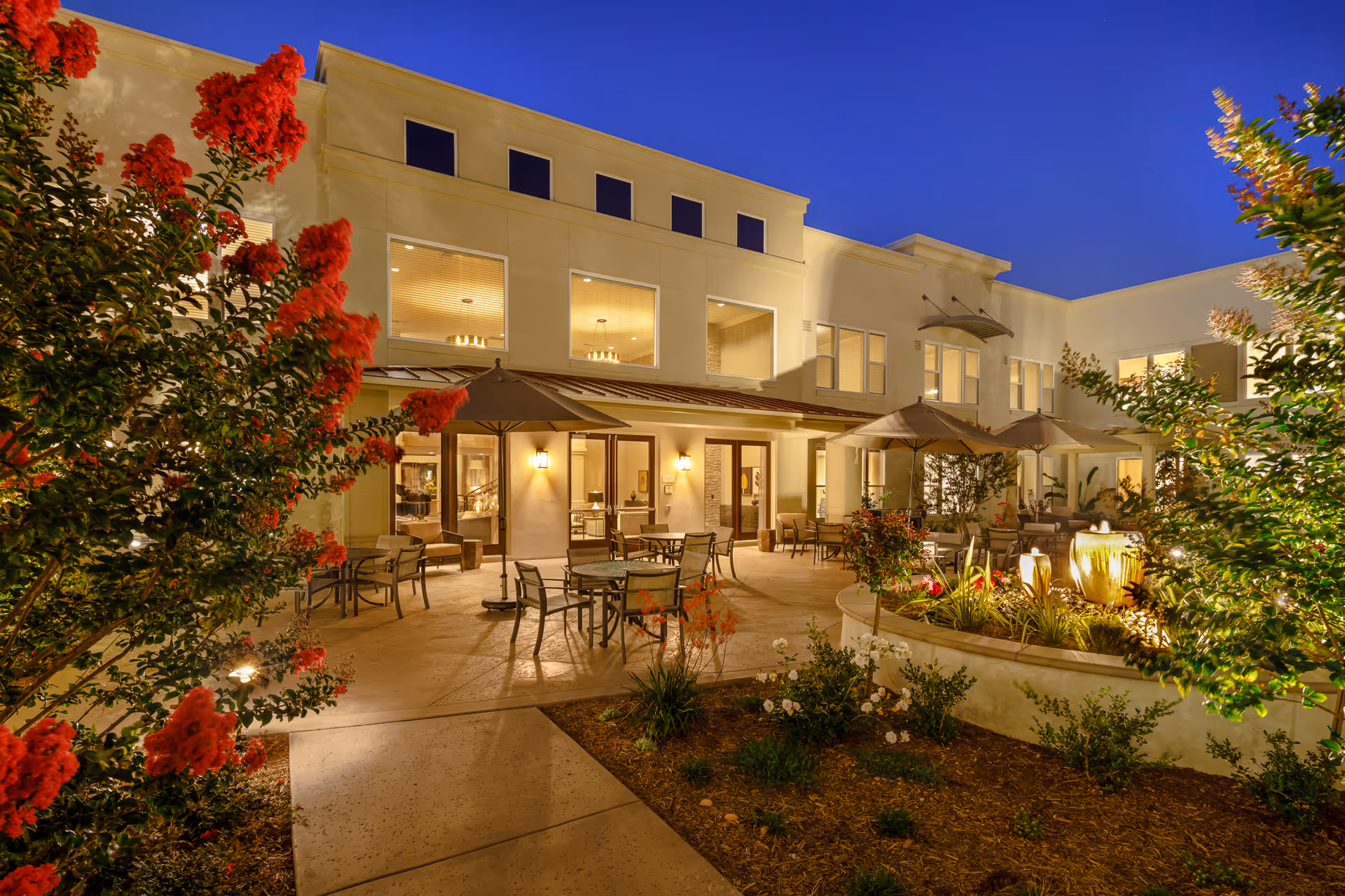A well-lit courtyard at dusk with patio tables and umbrellas, a small fountain, and a surrounding two-story building.