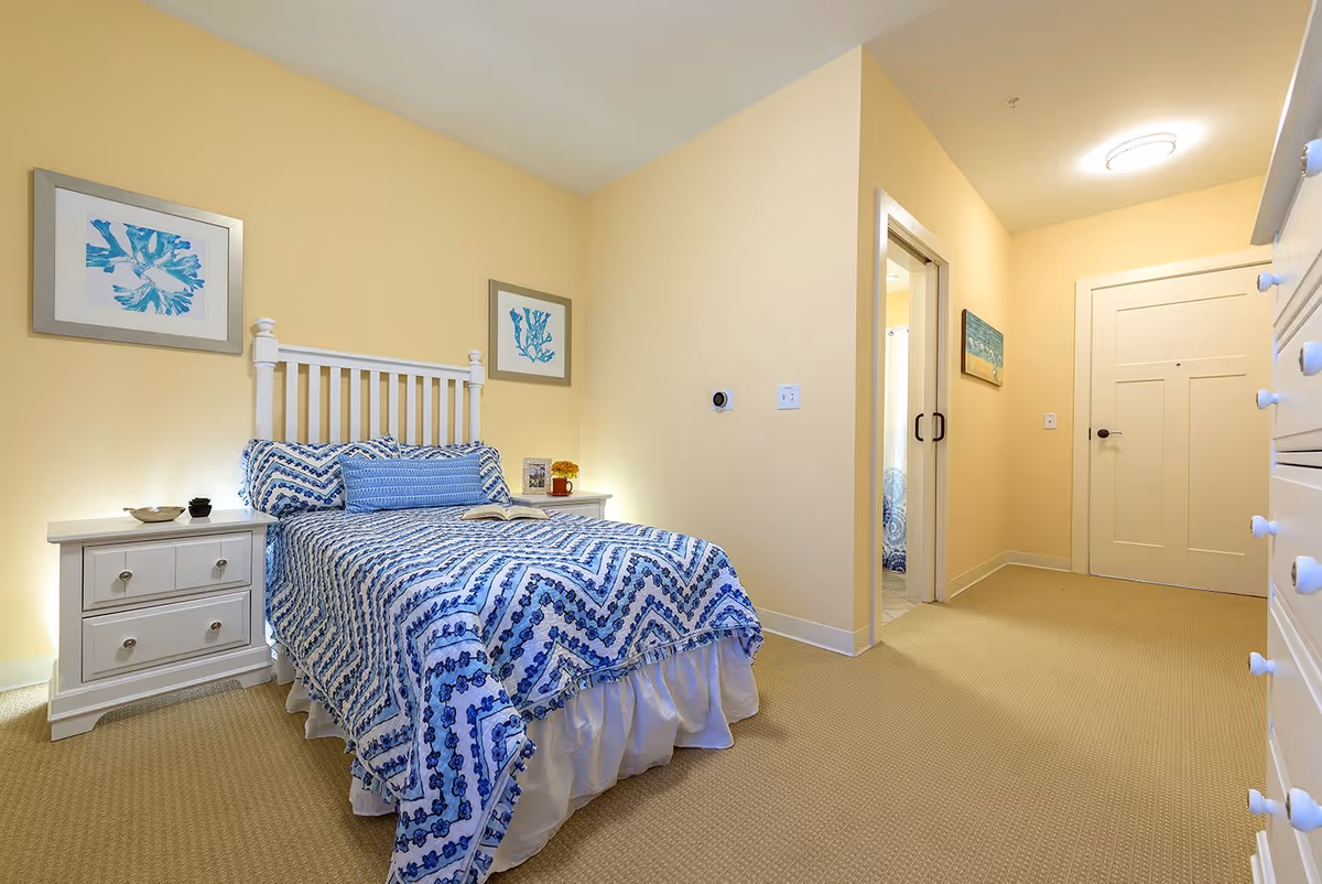 A cozy bedroom with a white wooden bed frame and blue patterned bedding. There are two framed artworks with blue designs on the light yellow walls above the bed. A white nightstand with drawers is beside the bed, holding a small plant and decorative items. The room has beige carpeting and a hallway leading to a closed white door and a bathroom with a sliding door.