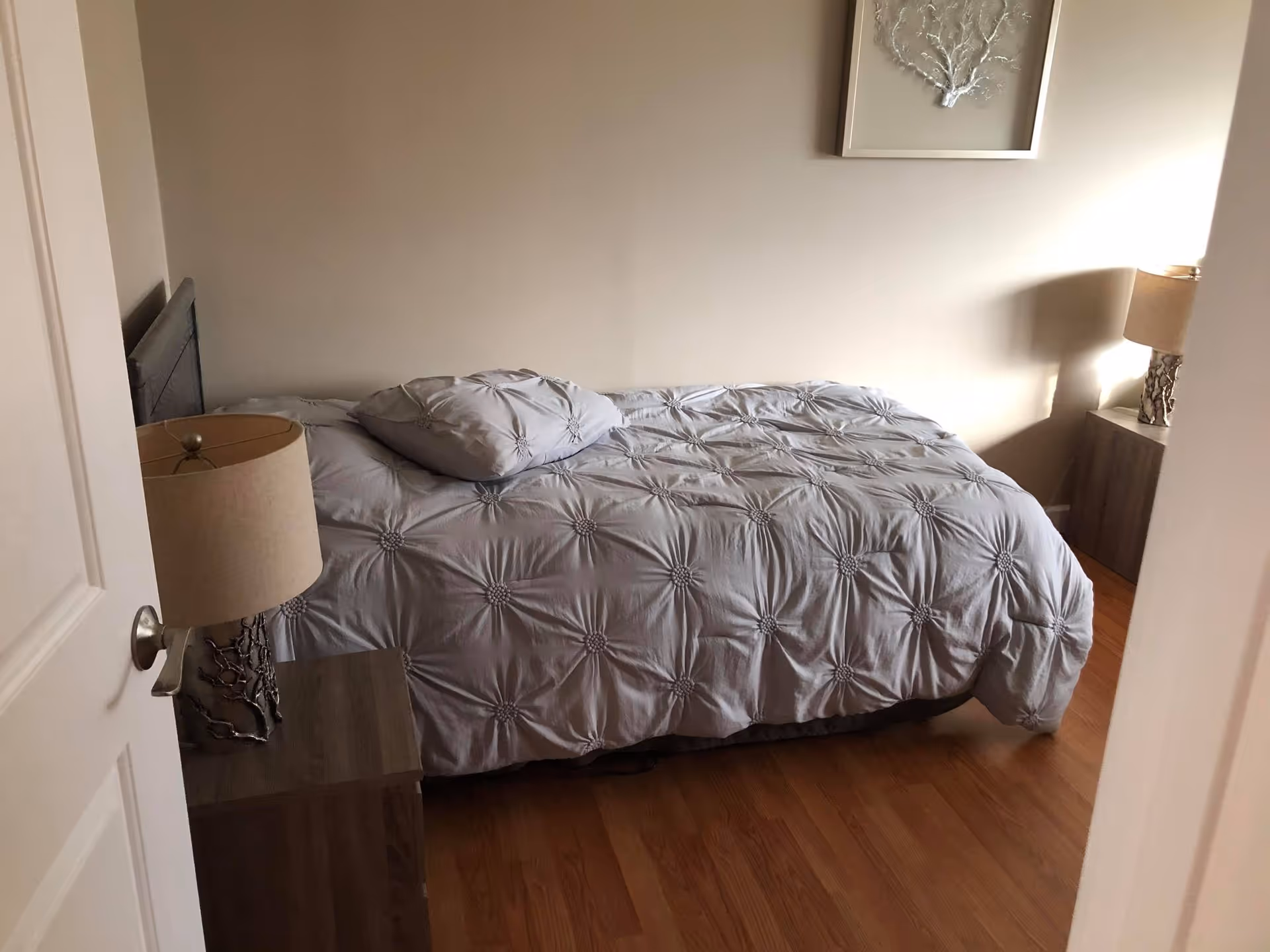 A small bedroom with a bed covered in a light gray comforter and a matching pillow. There are two wooden nightstands on either side of the bed, each with a lamp featuring a beige shade and a decorative base. The room has light-colored walls and wooden flooring, with a framed piece of wall art above the bed.