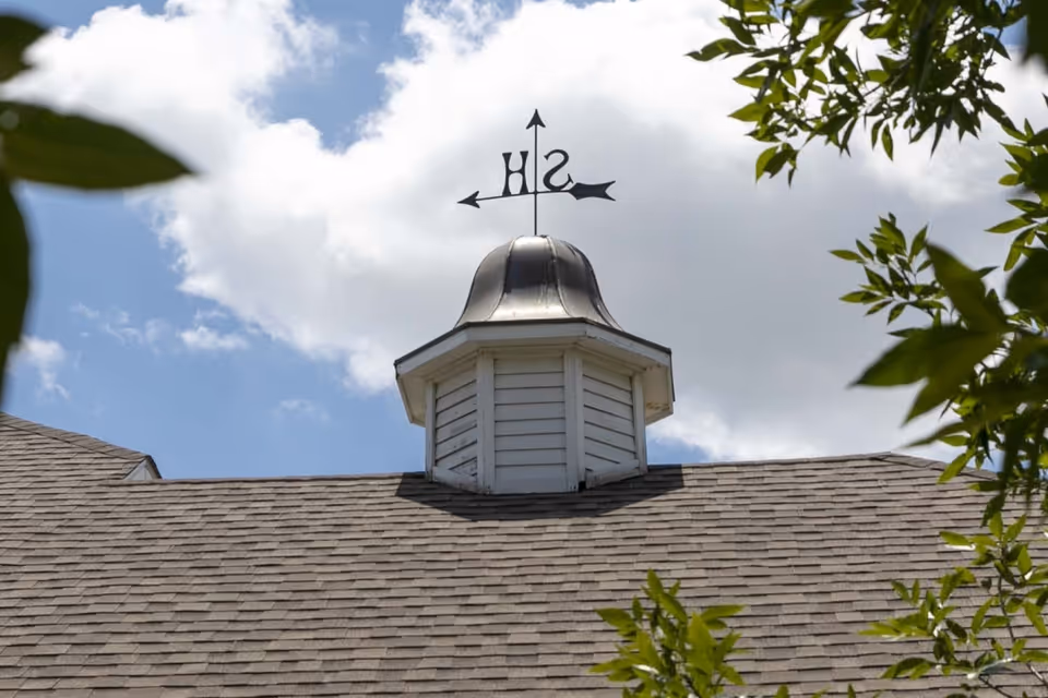 Close-up view of a rooftop cupola with a weather vane on top, showing directions S and H, under a partly cloudy sky with tree branches framing the image.