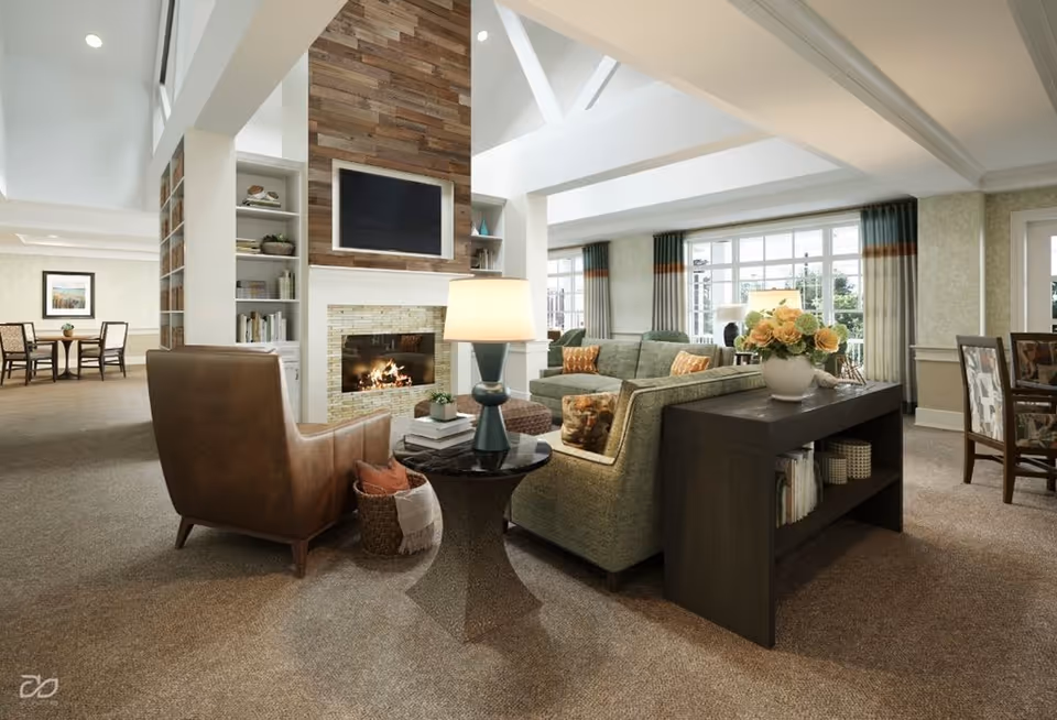 A cozy living room area in a senior living facility featuring a green sofa, a brown leather armchair, a side table with a lamp, and a fireplace with a mounted TV above it. The room has large windows with curtains, a bookshelf built into the fireplace wall, and a console table with a vase of flowers and books. In the background, there is a dining area with tables and chairs.