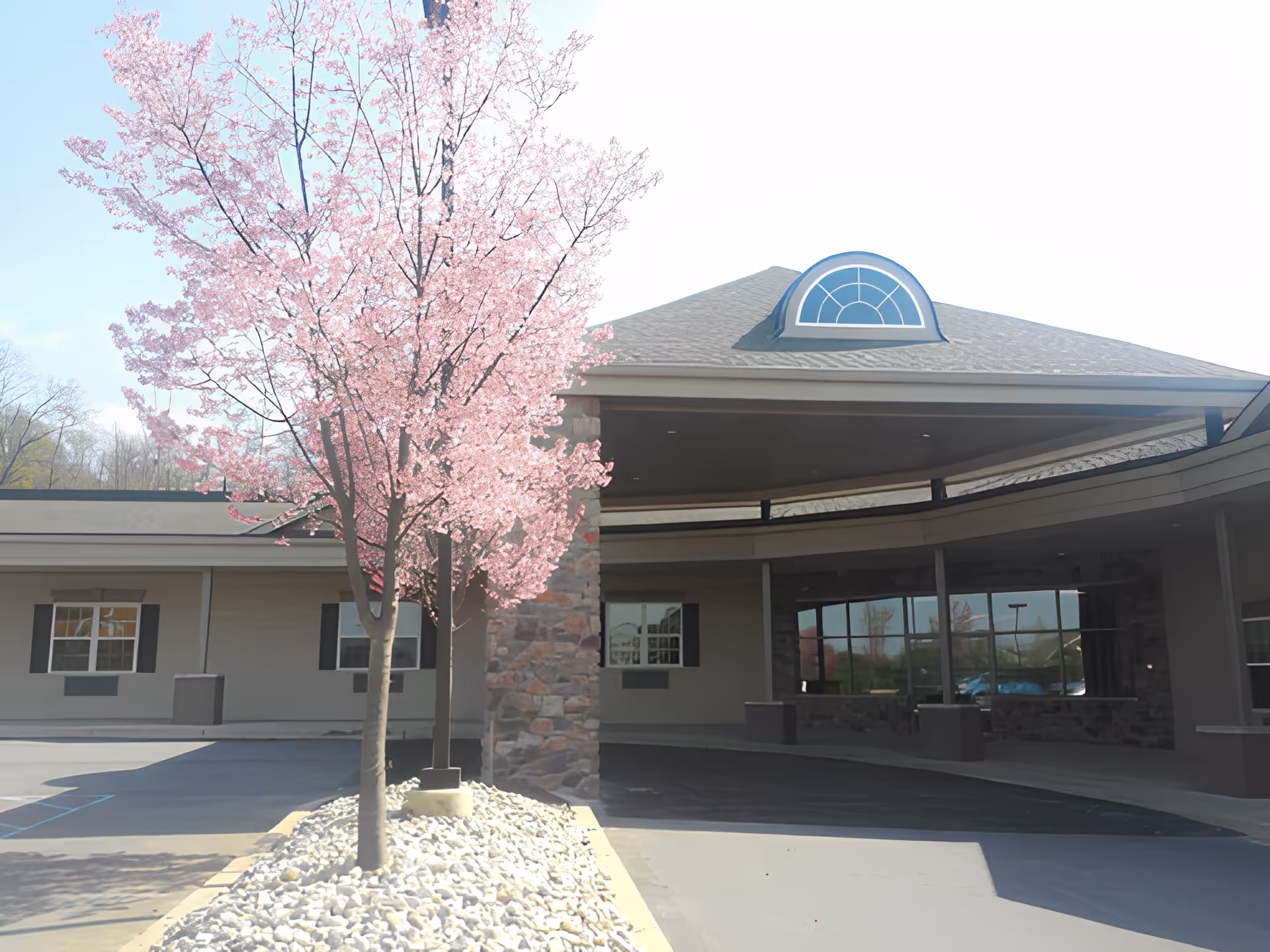 Exterior view of Abington Manor At Morgan Hill Memory Care Village showing the entrance with a covered driveway, a stone pillar, and a tree with pink blossoms in the foreground.