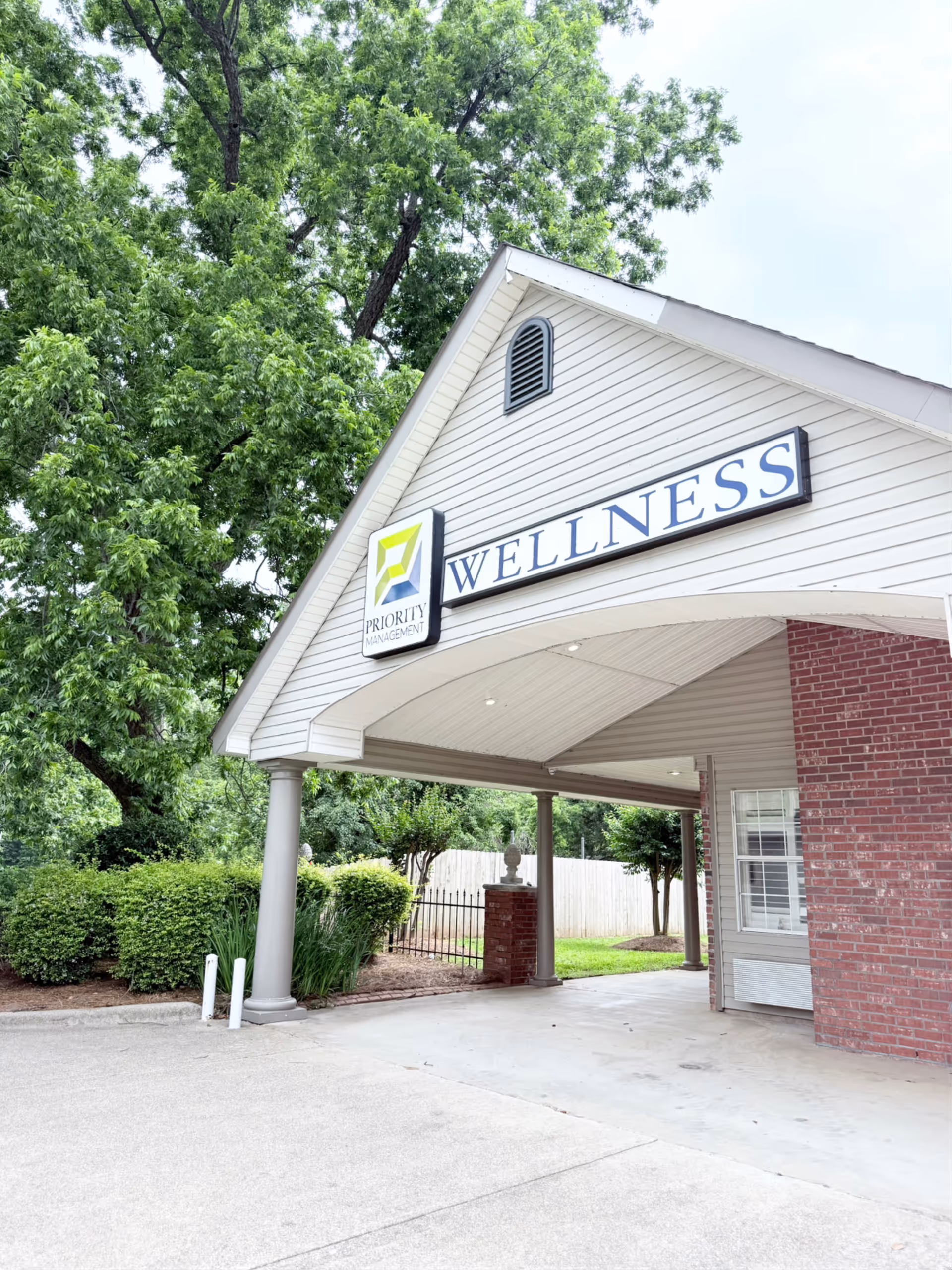 Entrance area of a building with a covered driveway supported by columns. The building has white siding and red brick walls. A sign above the entrance reads 'PRIORITY MANAGEMENT WELLNESS'. There are green trees and bushes surrounding the area.