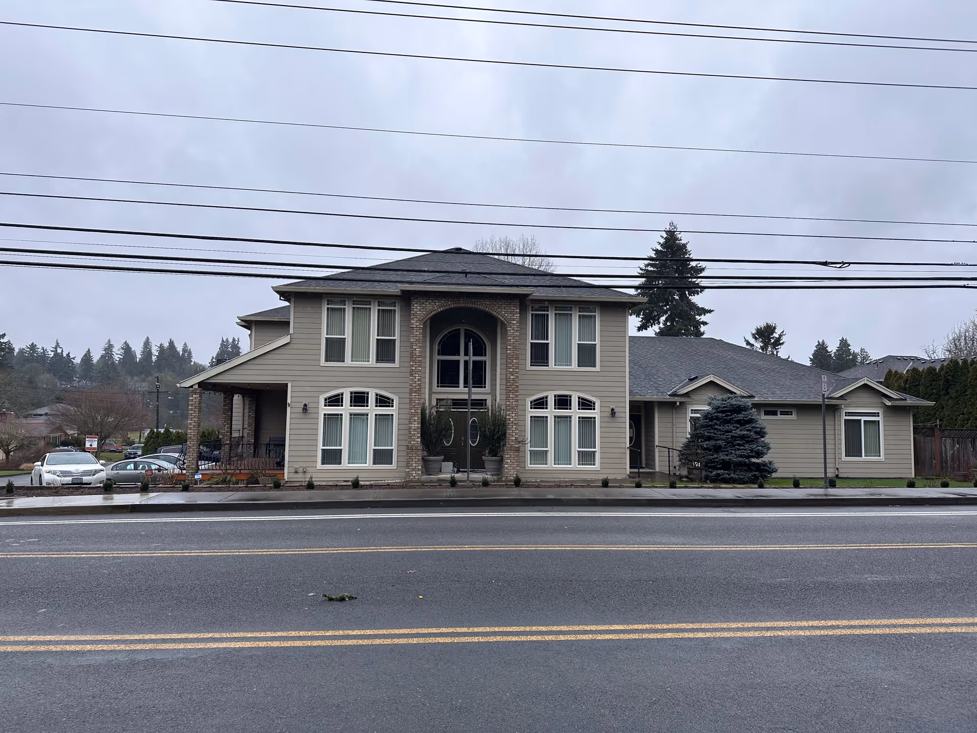 Exterior view of a two-story senior care facility building on a cloudy day. The building has beige siding with brick accents around the entrance, multiple windows, and a small landscaped area with shrubs and a tree. There are power lines visible in front of the building and a road with yellow dividing lines in the foreground.