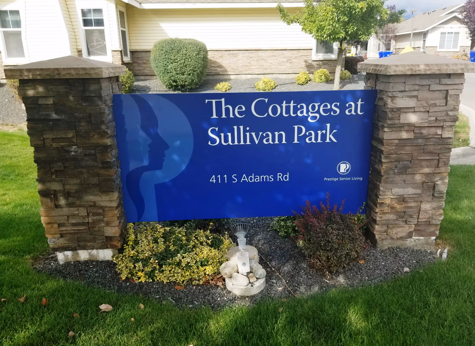 A blue sign with white text reading 'The Cottages at Sullivan Park, 411 S Adams Rd' is displayed between two stone pillars. The sign includes a logo and the text 'Prestige Senior Living'. The sign is surrounded by green grass, small bushes, and landscaping rocks. In the background, parts of residential buildings are visible.