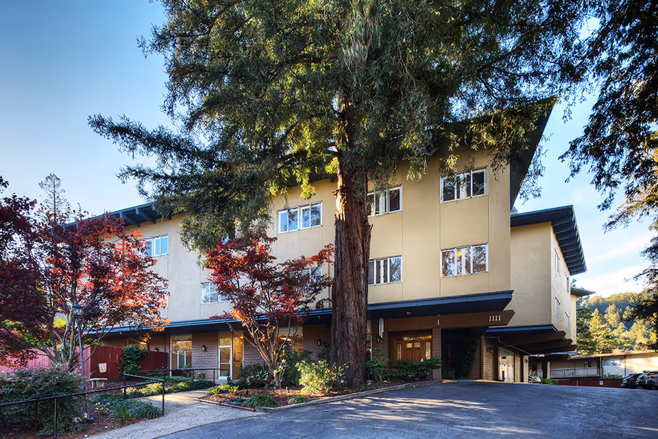 Exterior view of a three-story senior living facility building surrounded by trees and landscaping, with a driveway and entrance visible under a covered area.