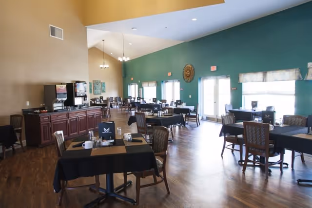 Spacious dining room with multiple set tables and chairs, a beverage station on the left, and high ceilings with large windows.