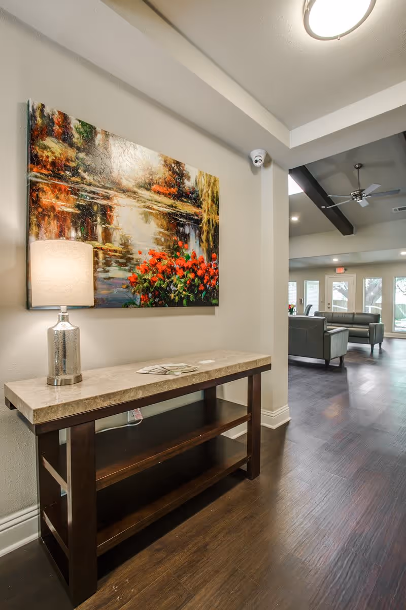 Interior view of a senior living facility showing a hallway with a wooden console table topped with a lamp and some brochures. Above the table is a colorful painting of a pond with red flowers. In the background, there is a common area with gray sofas, large windows, and a ceiling fan.