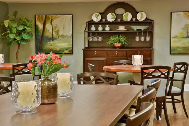Dining area with wooden tables and chairs, a decorative hutch, wall paintings, and floral centerpieces.