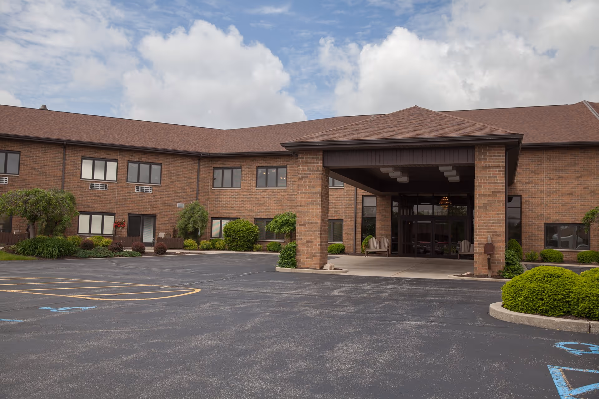 Brick two-story senior living building with a covered main entrance, landscaped shrubs, and an empty parking lot.