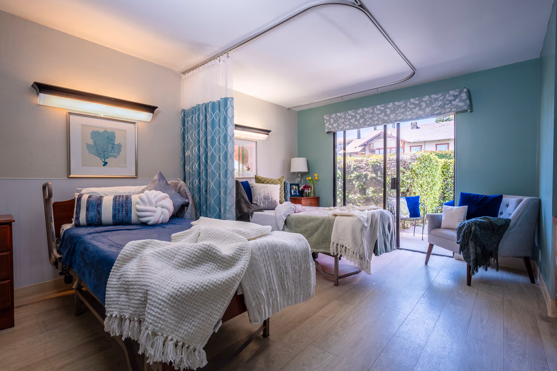 A bright and cozy bedroom in a senior living facility with two beds separated by a privacy curtain. Each bed is neatly made with blankets and decorative pillows. The room features soft lighting, framed artwork on the walls, a small wooden nightstand with a lamp and flowers, and a sliding glass door that opens to a garden area outside. There is also a comfortable armchair with a throw blanket near the door.