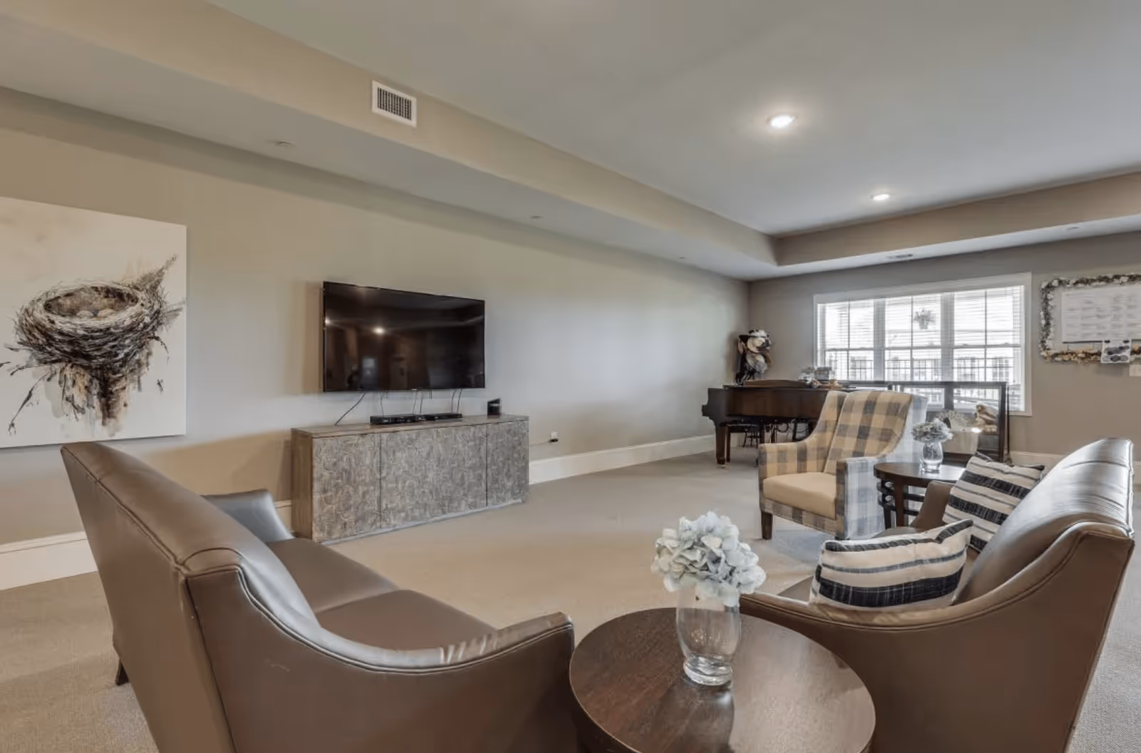 A spacious living room with beige walls and carpeted floor featuring a wall-mounted flat-screen TV above a decorative cabinet. The room includes two brown leather sofas with striped pillows, a round wooden coffee table with a glass vase of flowers, a plaid armchair, and a grand piano near a large window with blinds. A painting of a bird's nest hangs on the wall beside the TV.