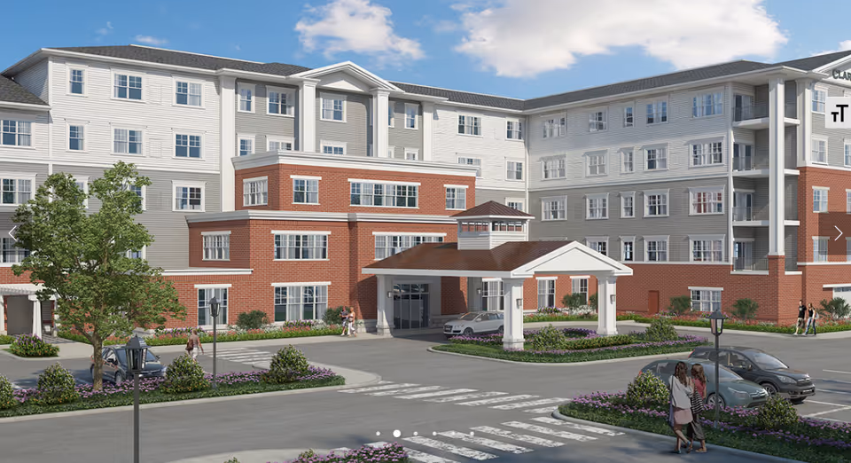 Front exterior of a multi-story senior living building with a covered entrance, driveway, landscaping, and parked cars.