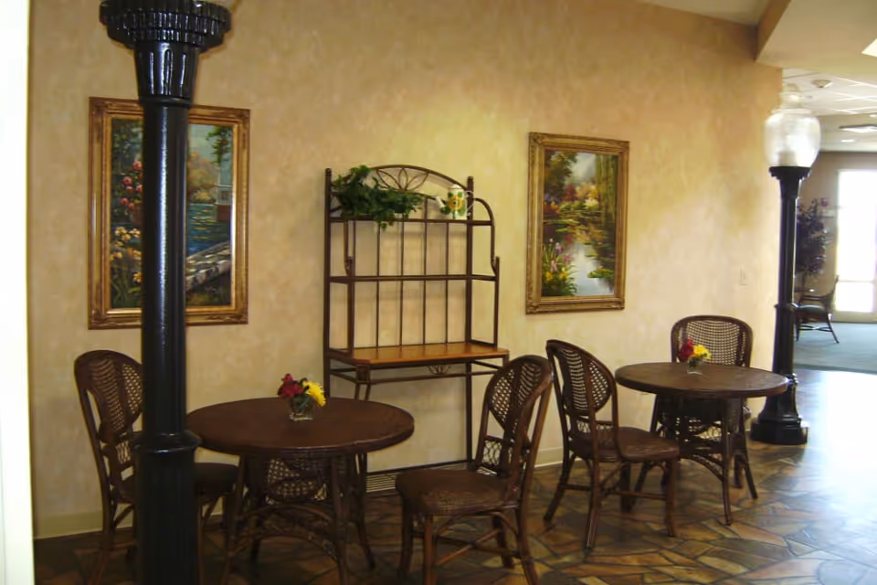Interior seating area with two round wooden tables, each surrounded by three wicker chairs. The walls are beige with two framed landscape paintings. A metal shelving unit with a plant and decorative items is against the wall. Two black lamp posts with glass light fixtures are positioned near the tables. The floor has a patterned tile design.