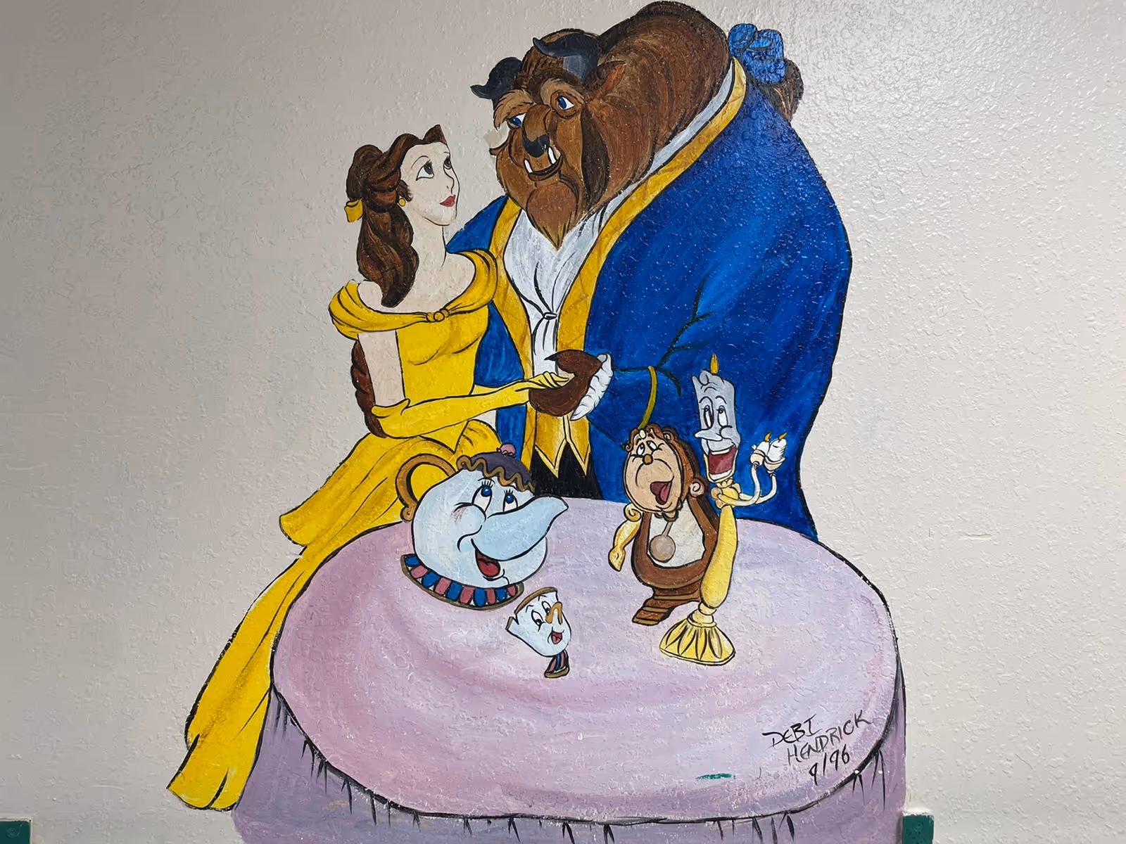 A colorful wall mural showing Belle and the Beast with Mrs. Potts, Chip, Lumière and Cogsworth above a pink table.