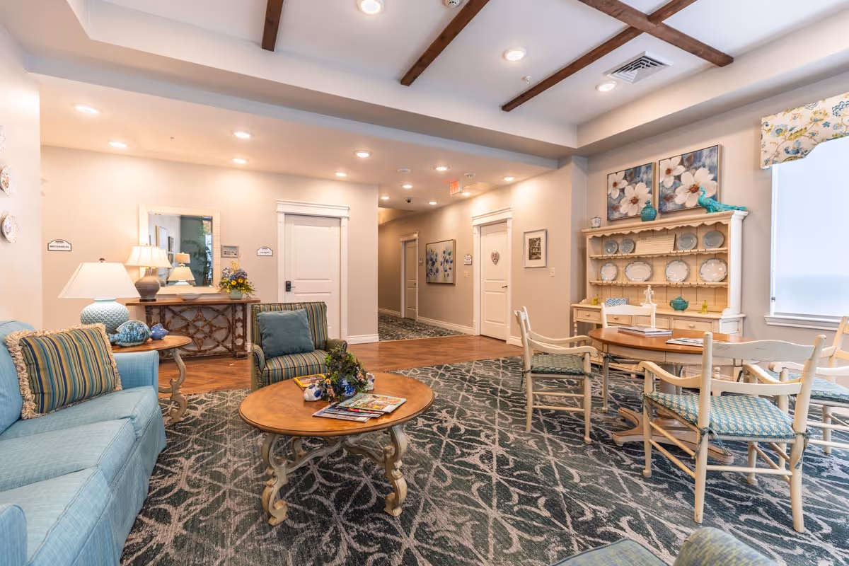 Bright, furnished common living area with sofas, chairs, a coffee table and a small dining table, decorative hutch and soft overhead lighting.