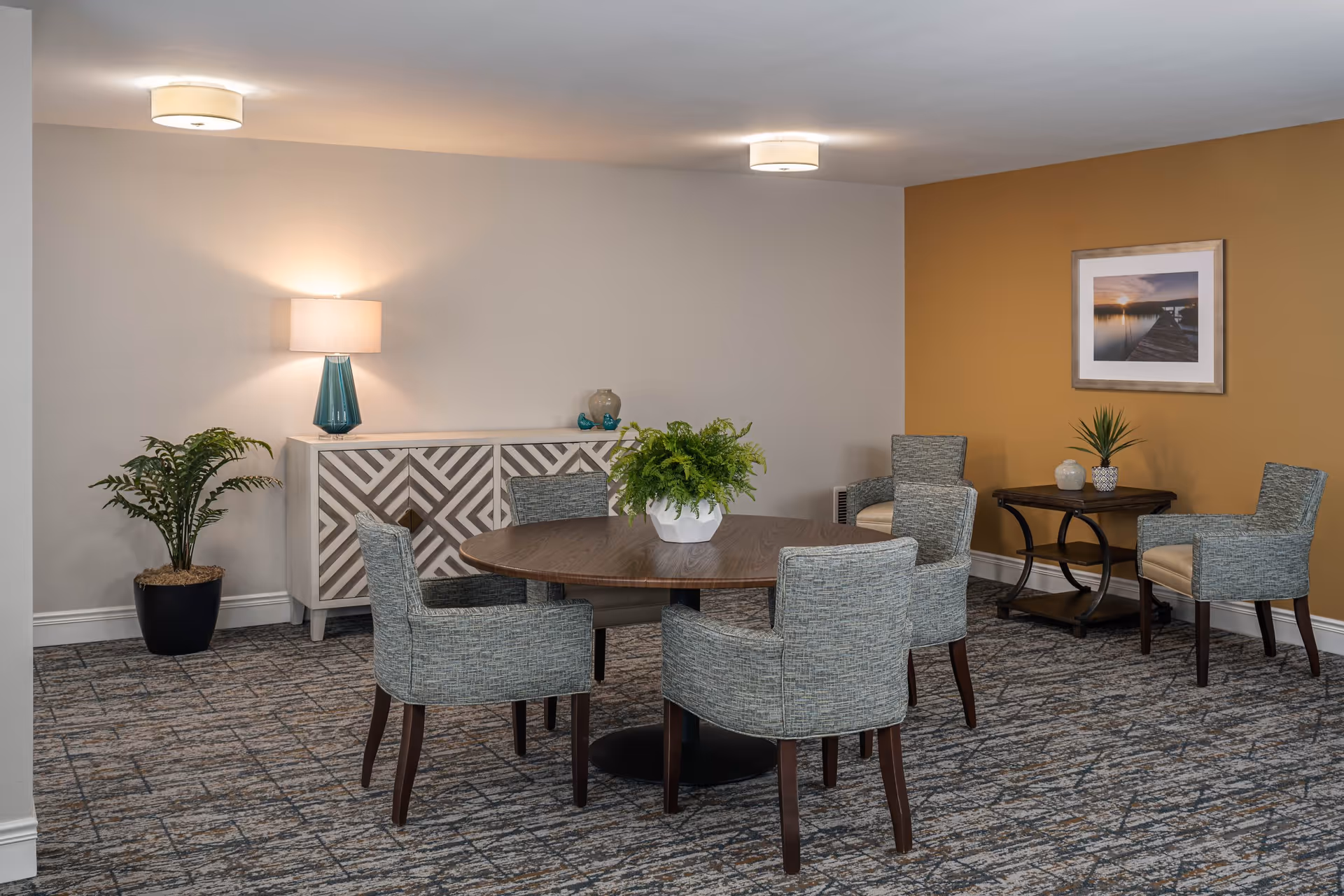 A round wooden table surrounded by upholstered chairs in a decorated senior living common/dining area with plants and artwork.