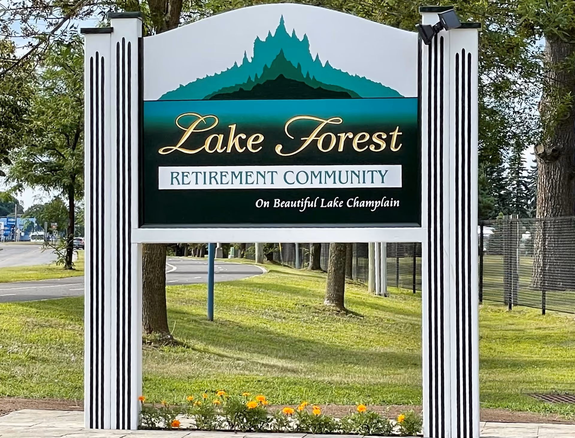 A large outdoor sign for Lake Forest Retirement Community, featuring a stylized green mountain graphic above the text. The sign is white with black vertical stripes on the supporting posts and is situated on a grassy area with a road and trees in the background.
