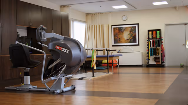 A spacious rehabilitation or physical therapy room with exercise equipment including a SciFit machine, parallel bars, and a rack holding various exercise bands and weights. The room has wood-patterned flooring, light-colored walls, a framed abstract painting, and a clock above a radiator cover.