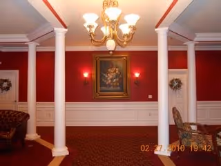 Interior view of a room with red walls and white wainscoting, featuring two white columns, a chandelier with multiple lights, a framed floral painting on the wall, and partial views of a brown leather sofa and a patterned chair.