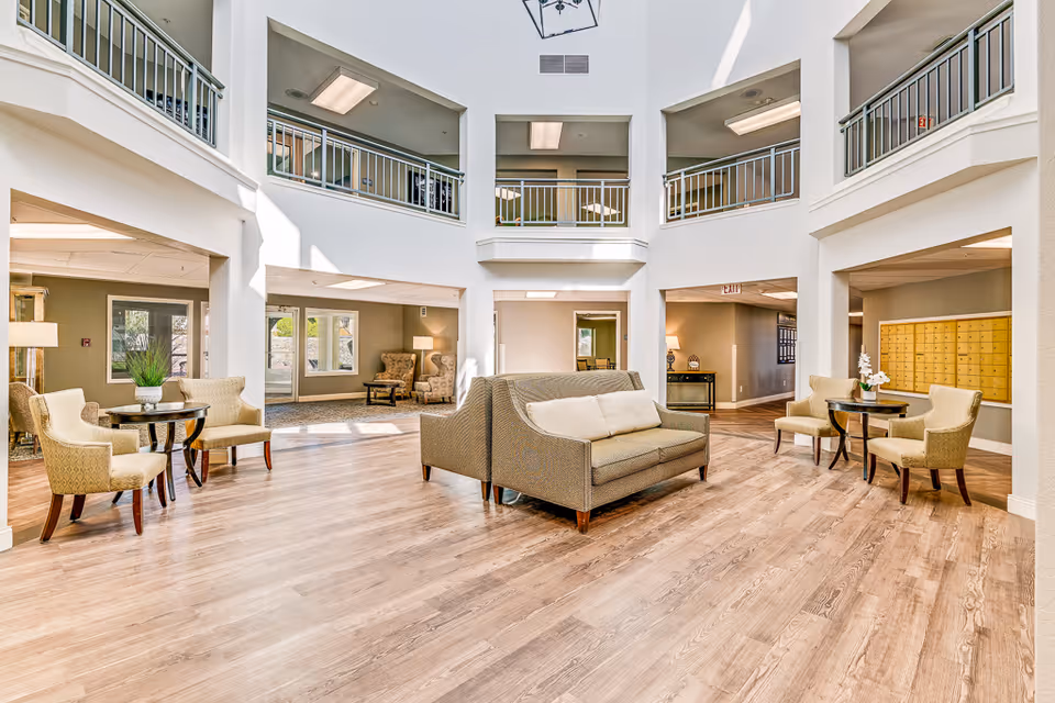Bright and spacious senior living facility common area with high ceilings and upper balcony railings. The room features light wood flooring, multiple seating areas with beige upholstered chairs and sofas, small round tables with plants and flowers, and large windows letting in natural light. There are mailboxes on one wall and various lamps providing additional lighting.